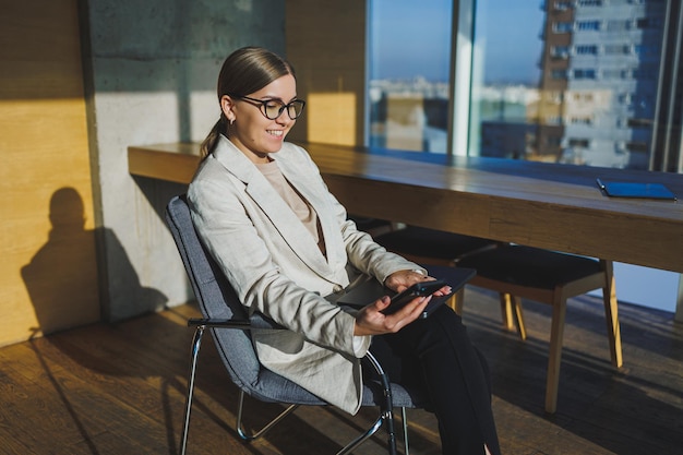 Trabalhe em um escritório moderno Uma jovem empresária positiva em roupas casuais e óculos, sentada no escritório com um telefone celular enquanto trabalha em um projeto remoto usando um laptop