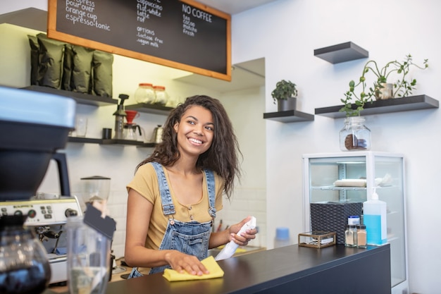 Trabalho, café. Jovem alegre, morena, com longos cabelos escuros em pé olhando para o lado, limpando o balcão do bar