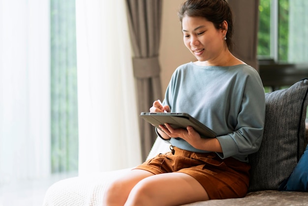 Trabalho casual em casa relaxe casual usando dispositivo tablet na sala de estar mulher adulta asiática digitando pesquisa ou mensagens de texto via sofá de laptop trabalhando em casa com o conceito de idéias sorridentes de felicidade pacífica