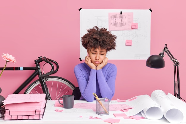 Trabalho de casa chato. triste insatisfeita jovem e ocupada afro-americana se sente cansada de poses rotineiras de trabalho na área de trabalho cercada de plantas, esboços, xícara de café