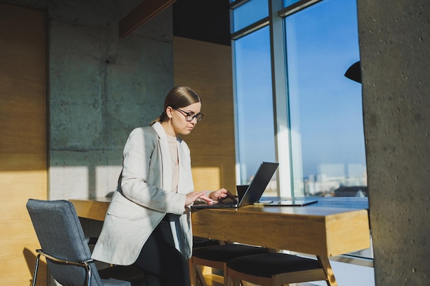 Trabalho de escritório atraente jovem gerente feminina trabalhando no escritório em pé no espaço de trabalho brilhante usando computador portátil moderno jovem bebendo café delicioso