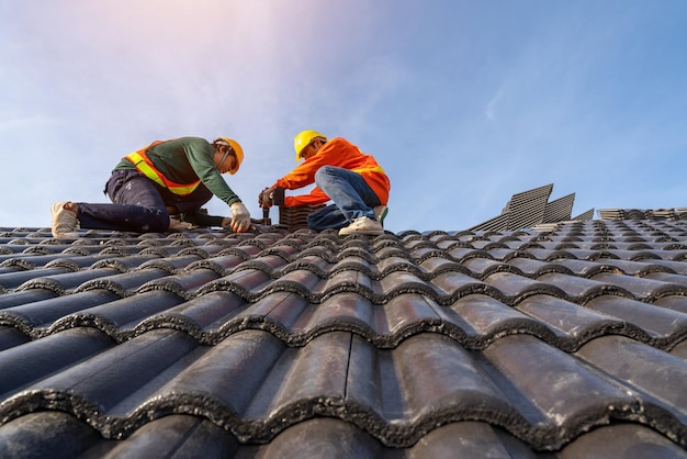 Trabalho em equipe de trabalhadores de telhados asiáticos em uniformes de proteção e luvas usando pistola de pregos pneumática ou pneumática e instalando telhas de concreto no topo do novo edifício do telhado em construção