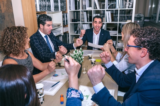 Trabalho em equipe feliz comemorando o sucesso do negócio com as mãos ao alto, sentado à mesa na sede da empresa