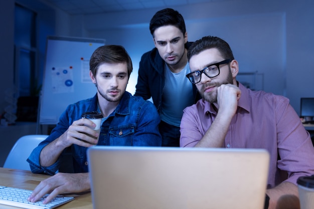 Foto trabalho em equipe. homem bonito e inteligente pensativo olhando para a tela do laptop e pensando em seu projeto enquanto trabalham juntos