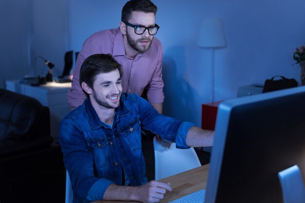 Trabalho favorito. Programador homem alegre e encantado sorrindo e apontando para a tela enquanto mostra algo para seu colega