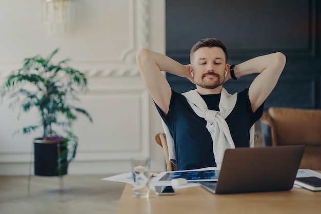 Trabalho feito. freelancer ou empresário relaxado em fones de ouvido sem fio sentado à mesa com as mãos atrás da cabeça, assistindo a webinar de negócios ou ouvindo alto-falante no laptop enquanto trabalha remotamente de casa