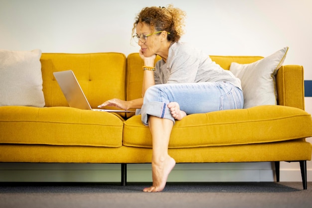 Trabalho freelancer jovem feliz no computador digitando mensagens de texto do conceito de tecnologia doméstica Uma pessoa do sexo feminino usando laptop no sofá amarelo e sorrindo Atividade de navegação na web Vista lateral Interior