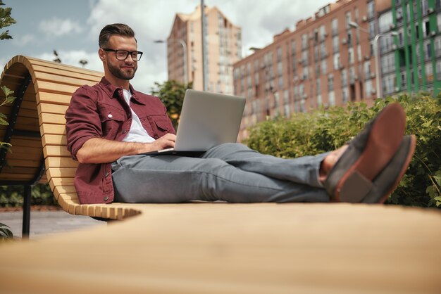 Trabalho perfeito. Homem bonito jovem feliz com barba por fazer em roupas casuais e óculos, trabalhando no laptop e sorrindo enquanto está sentado no banco ao ar livre. Blogging. Vida urbana