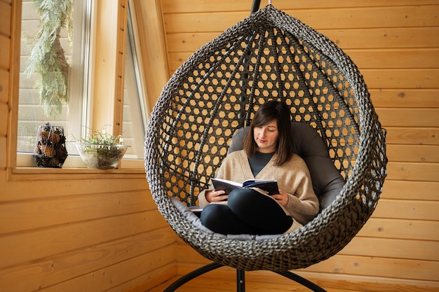 Trabalho remoto e fuga para o conceito de natureza A mulher senta-se no balanço da cadeira do ovo e lê o livro na pequena cabana de madeira