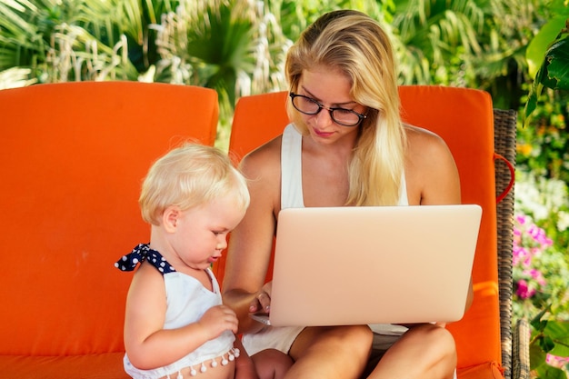 Trabalho remoto e surf freelancer jovem mãe empresária freelancer trabalhando com laptop sentado em uma espreguiçadeira ao lado de sua filha bebê licença maternidade em um resort tropical praia de verão
