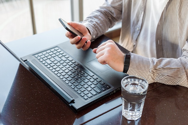 Trabalho remoto. Homem usando laptop, segurando o telefone inteligente para negócios trabalho ou estudo, relógio inteligente. Espaço de trabalho freelancer, empregado em teletrabalho. Feche acima do espaço de trabalho remoto em casa, coworking, café, escritório.