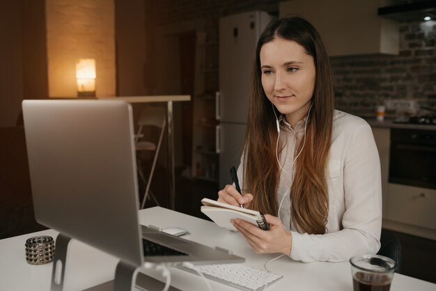 Trabalho remoto. Uma mulher caucasiana com fones de ouvido trabalhando remotamente em seu laptop. Uma garota em uma camisa branca, fazendo anotações durante uma reunião de negócios on-line em seu local de trabalho em casa acolhedor.