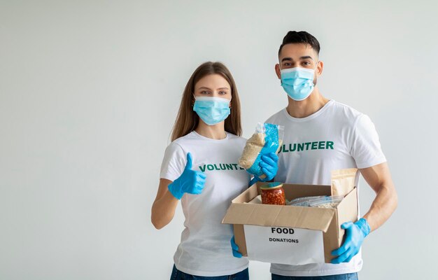 Trabalho voluntário jovens voluntários felizes em máscaras médicas segurando caixa de doações de comida mulher mostrando o polegar para cima o espaço da cópia