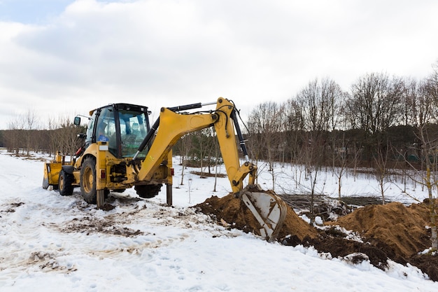 Un tractor excavador amarillo excava el suelo para la reparación y el tendido de servicios públicos subterráneos y cables para electricidad y comunicaciones. Durante la temporada de invierno. Foto de alta calidad