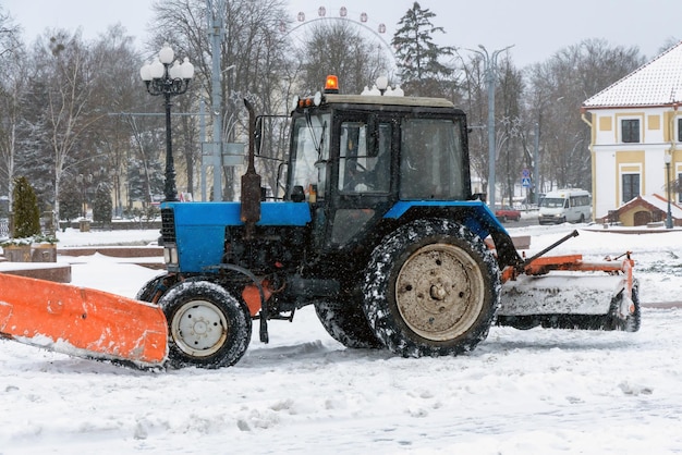 Un tractor limpia la nieve en la ciudad en invierno después de una nevada Limpiando las calles de la ciudad de la nieve Conductor del tractor en el trabajo en la plaza de la ciudad durante una tormenta de nieve