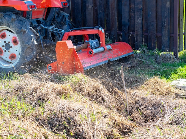 Un tractor con una podadora colocada cubre pasto seco a lo largo de la cerca. Procesamiento de parcelas de tierra