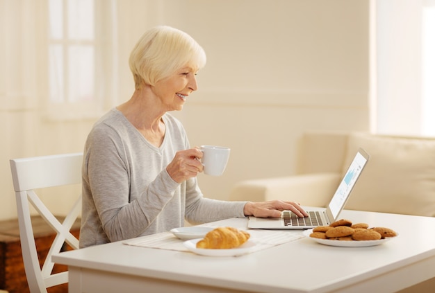 Tradição matinal. Mulher madura alegre sentada em posição semi-aberta e segurando o copo com a mão direita enquanto usa seu laptop