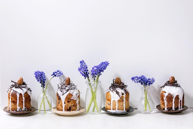 Tradición casera, pequeños pasteles kulich de Pascua con nidos de chocolate y huevos en platos en fila decorados con flores de muscari sobre una mesa de mármol blanco. Horneado tradicional ortodoxo de Pascua ruso ucraniano