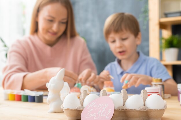 Tradición familiar antes de Pascua: centrarse en huevos en cartón y tarjeta de Pascua rosa, madre e hijo pintando huevos