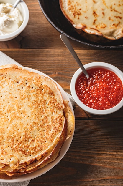 Tradicional crepes russos blini empilhados no prato e frigideira de ferro fundido com caviar vermelho, creme de leite fresco na mesa de madeira escura. refeição festiva russa maslenitsa ou entrudo.