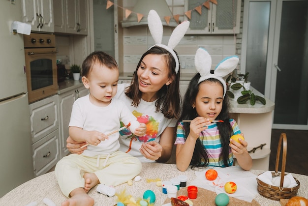 Tradiciones familiares de Pascua Amante joven madre enseñando a los niños a pintar huevos para Pascua mientras se sientan juntos en la mesa de la cocina