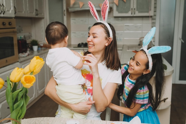 Tradiciones familiares de Pascua Amante joven madre enseñando a los niños a pintar huevos para Pascua mientras se sientan juntos en la mesa de la cocina