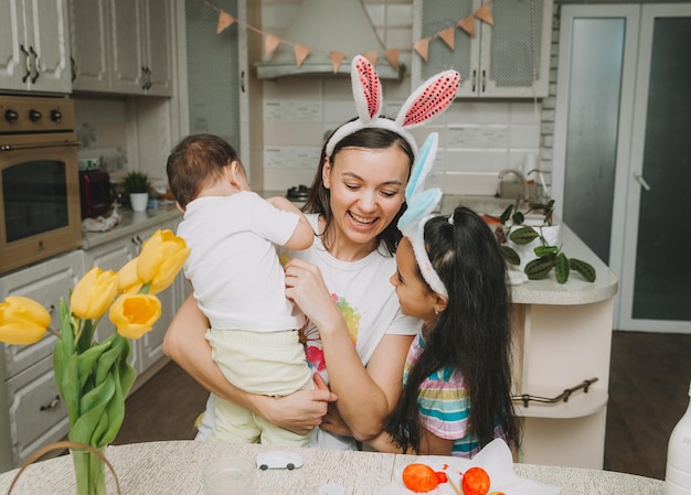 Tradiciones familiares de Pascua Amante joven madre enseñando a los niños a pintar huevos para Pascua mientras se sientan juntos en la mesa de la cocina