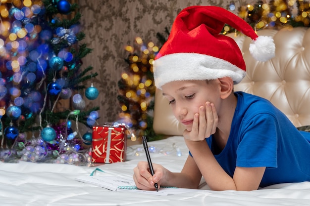 Tradições de ano novo e natal. um menino caucasiano com um chapéu de papai noel vermelho escrevendo uma carta para o papai noel em casa