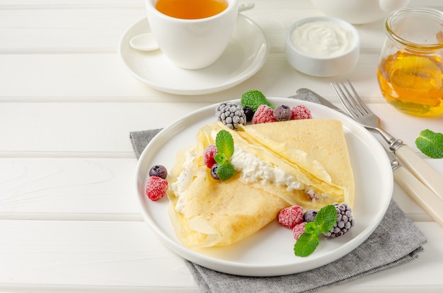 Traditionelle dünne Pfannkuchen oder Crepes mit Hüttenkäse und Rosinen mit Beeren, Honig und Sauerrahm auf einem weißen hölzernen Hintergrund. Essen für Maslenitsa. Speicherplatz kopieren.