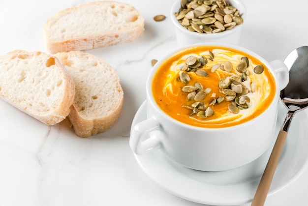 Traditionelle Herbst- und Winterteller, heiße und würzige Kürbissuppe mit Kürbiskernen, Creme und frisch gebackenem Stangenbrot, auf weißer Marmortabelle, Kopienraum