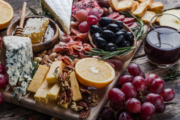 Traditionelle italienische Antipasti-Platte. Verschiedene Käsesorten auf Holzschneidebrett. Brie-Käse, Cheddar-Scheiben, Gogonzola, Walnuss-Trauben, Oliven, Schinken, Rosmarin und ein Glas Rotwein. Draufsicht