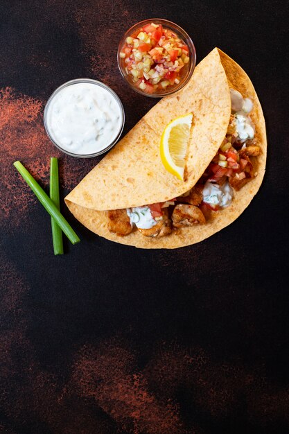 Traditionelle mexikanische Fajitas mit Hühnchen und Gemüse, serviert in Tortilla mit weißer und roter Sauce, Zitrone und frischen Frühlingszwiebeln. Draufsicht. dunkler Hintergrund. Speicherplatz kopieren