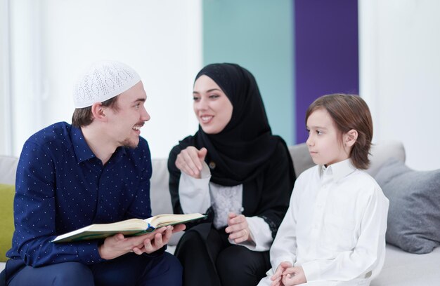 Traditionelle muslimische familieneltern mit kindern, die vor dem iftar-abendessen während eines ramadan-festes zu hause den koran lesen und gemeinsam auf dem sofa beten