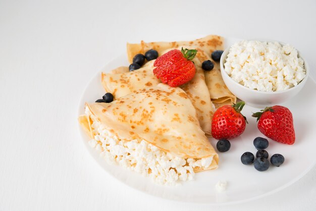 Traditionelle russische dünne Pfannkuchen mit Hüttenkäse und Beeren in einem Teller auf einem weißen, Kopierraum