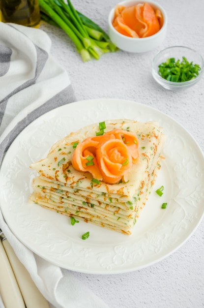 Traditionelle russische dünne Pfannkuchen oder Crepes mit Käse, Kräutern und geräuchertem Lachs auf einem weißen Teller auf hellem Hintergrund. Urlaub maslenitsa. Vertikale Ausrichtung.