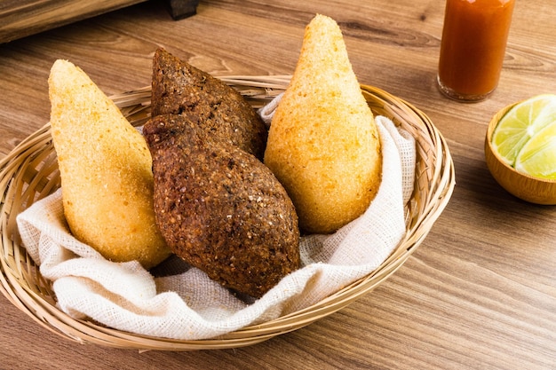 Traditionelle Snacks Chicken Coxinha, bekannt als Coxinha in Brasilien und Fried Kibe, serviert in einem Korb mit Gewürzen wie Zitrone und Pfeffer auf der Seite Holztisch Selektiver Fokus