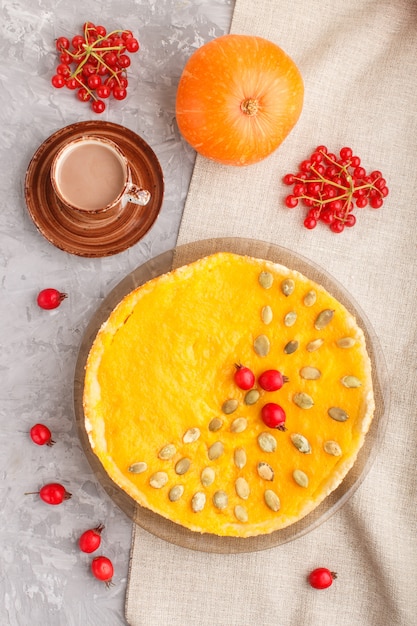 Traditioneller amerikanischer süßer Kürbiskuchen verziert mit roten Beeren und Kürbiskernen des Weißdorns mit Tasse Kaffee auf einem grauen Beton. Draufsicht, Nahaufnahme.