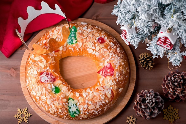 Traditioneller Dreikönigskuchen Roscon de Reyes mit Papierkrone auf Holztischplatte mit kleinem Weihnachtsbaum und Dekoration, Nahaufnahme, Ansicht von oben