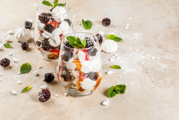 Traditioneller englischer Nachtisch. Eton Mess Schlagsahne, Baiser, frische Brombeeren, Sauce und Karamell. In Serviergläsern auf einem hellen Steintisch.