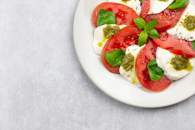 Traditioneller italienischer Caprese-Salat mit geschnittenen Tomaten, Mozzarella, Basilikum, Pesto-Sauce und Gewürzen auf hellgrauem Betonhintergrund. Ansicht von oben. Platz kopieren.