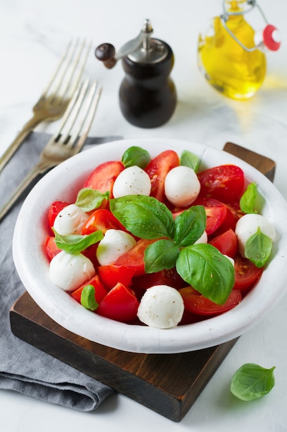 Traditioneller italienischer Caprese-Salat mit Tomaten, Maozzarella-Käse und Basilikum auf hellem Marmorhintergrund in einer weißen alten Keramikplatte.