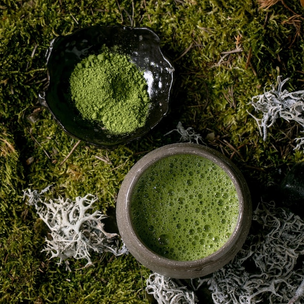 Traditioneller japanischer heißer grüner schaumiger Tee Matcha in Keramiktasse und Matcha-Pulver über Waldmoos als Hintergrund Gesundes natürliches Getränk Flaches quadratisches Bild