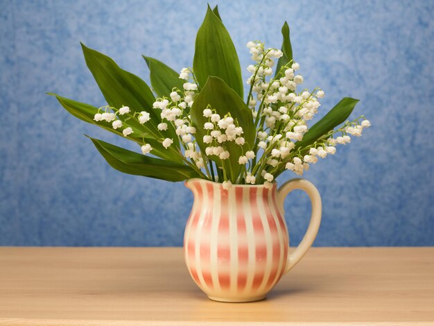 Traditioneller Krug mit Lilly of the Valley auf einem Sideboard, blaue Tapete im Hintergrund