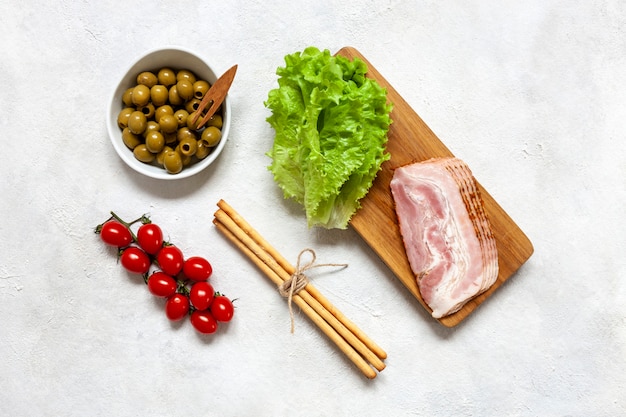 Traditioneller Pancetta-Speck, serviert mit eingelegten Oliven, Tomaten, grünen Salatblättern und Grissini-Brot