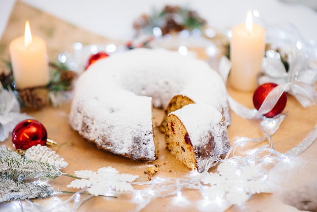 Traditioneller runder Christstollen aus getrockneten Früchten und Nüssen, bestreut mit Puderzucker auf dem Hintergrund eines Weihnachtsdekors mit Kerzen. Traditioneller Weihnachtskuchen.