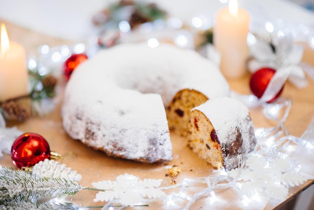 Traditioneller runder Christstollen aus getrockneten Früchten und Nüssen, bestreut mit Puderzucker auf dem Hintergrund eines Weihnachtsdekors mit Kerzen. Traditioneller Weihnachtskuchen.