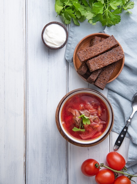 Traditioneller ukrainischer russischer Borschtsch (Rote-Bete-Suppe) mit Fleisch in einer schwarzen Schüssel auf einem hölzernen Hintergrund mit Scheiben gebratenem Brot, Sauerrahm und frischen Kräutern. Draufsicht und Platzbild kopieren