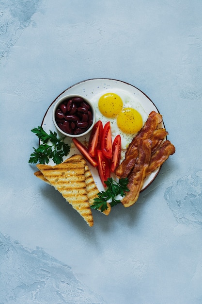 Traditionelles englisches Frühstück mit Spiegeleiern, Speck, Bohnen, Toast und Tomaten auf grauem Beton