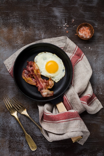 Traditionelles englisches Frühstück mit Spiegeleiern und Speck in einer gusseisernen Pfanne auf dunklem Betonhintergrund. Ansicht von oben.