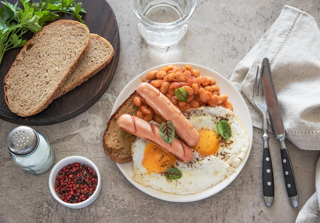 Traditionelles englisches Frühstück mit Spiegeleiern, Würstchen, Bohnen, frischem Kraut, rotem Pfeffer und Toast auf grauem Hintergrund. Konzeptmorgen und Nahrungsnahrung. Ansicht von oben.